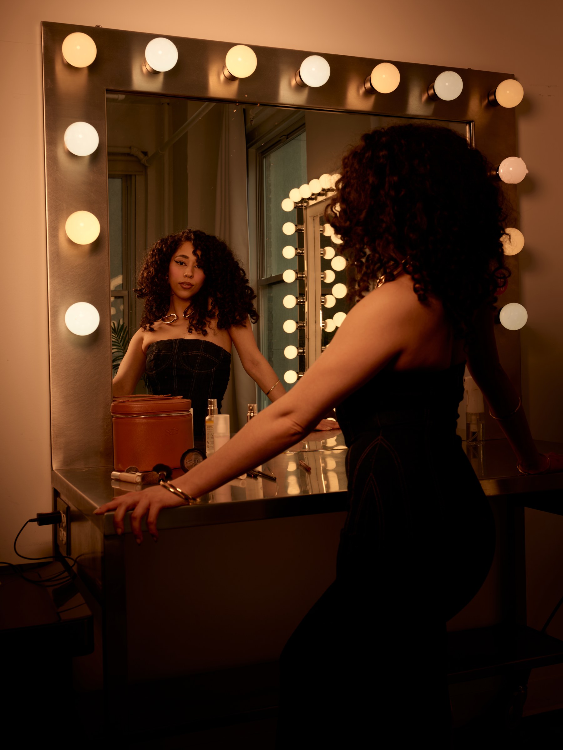 Woman seated at a lighted dressing-room mirror.