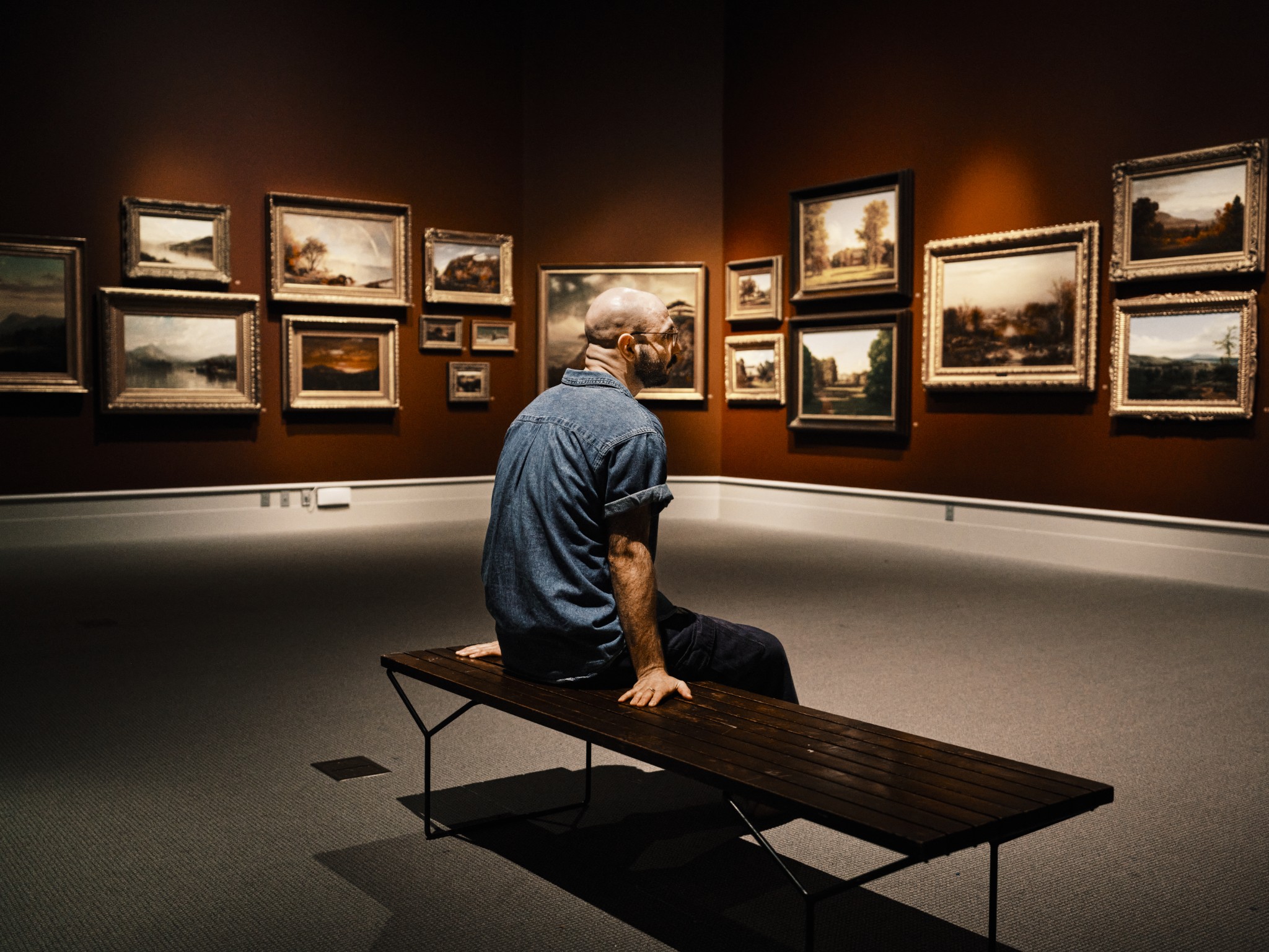 Man seated in a museum gallery surrounded by landscape paintings.